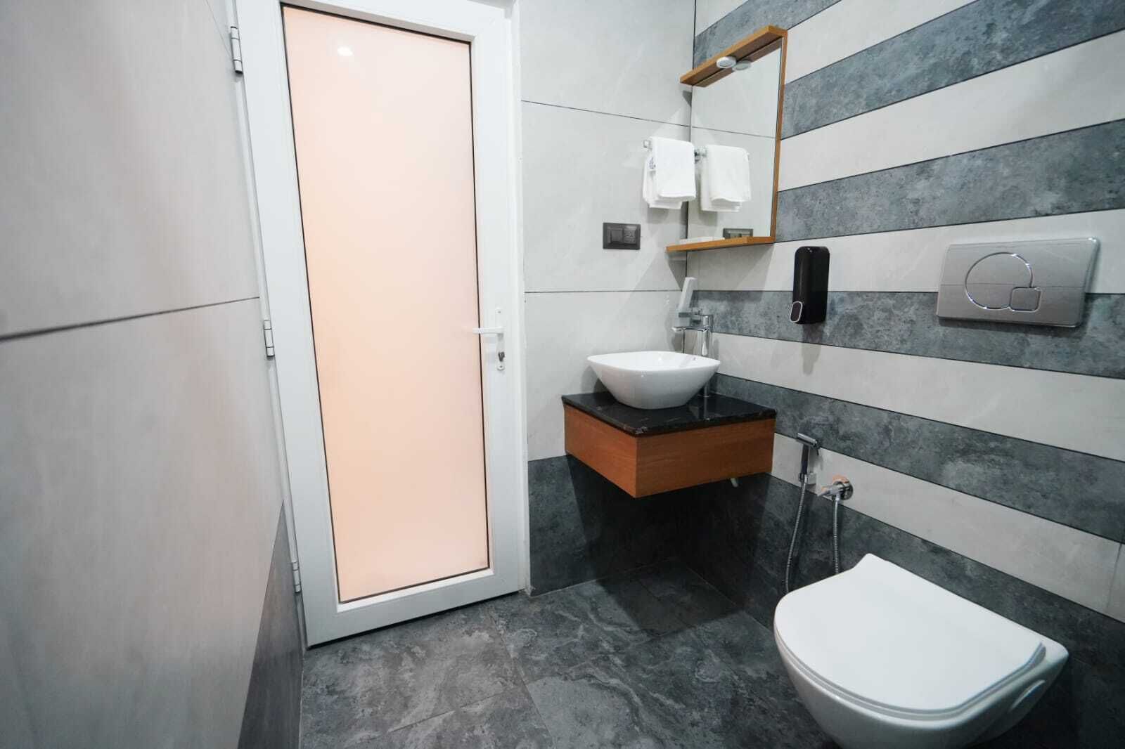 A modern bathroom with a white toilet, a white sink with a wooden base, a towel rack, a mirror, and a shower control panel.