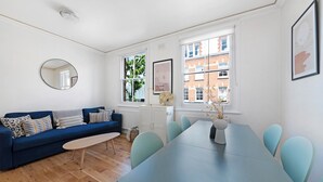 Ein modernes Wohnzimmer mit einem blauen Sofa, einem runden Spiegel, einem Esstisch mit Stühlen und großen Fenstern mit Blick auf Gebäude.