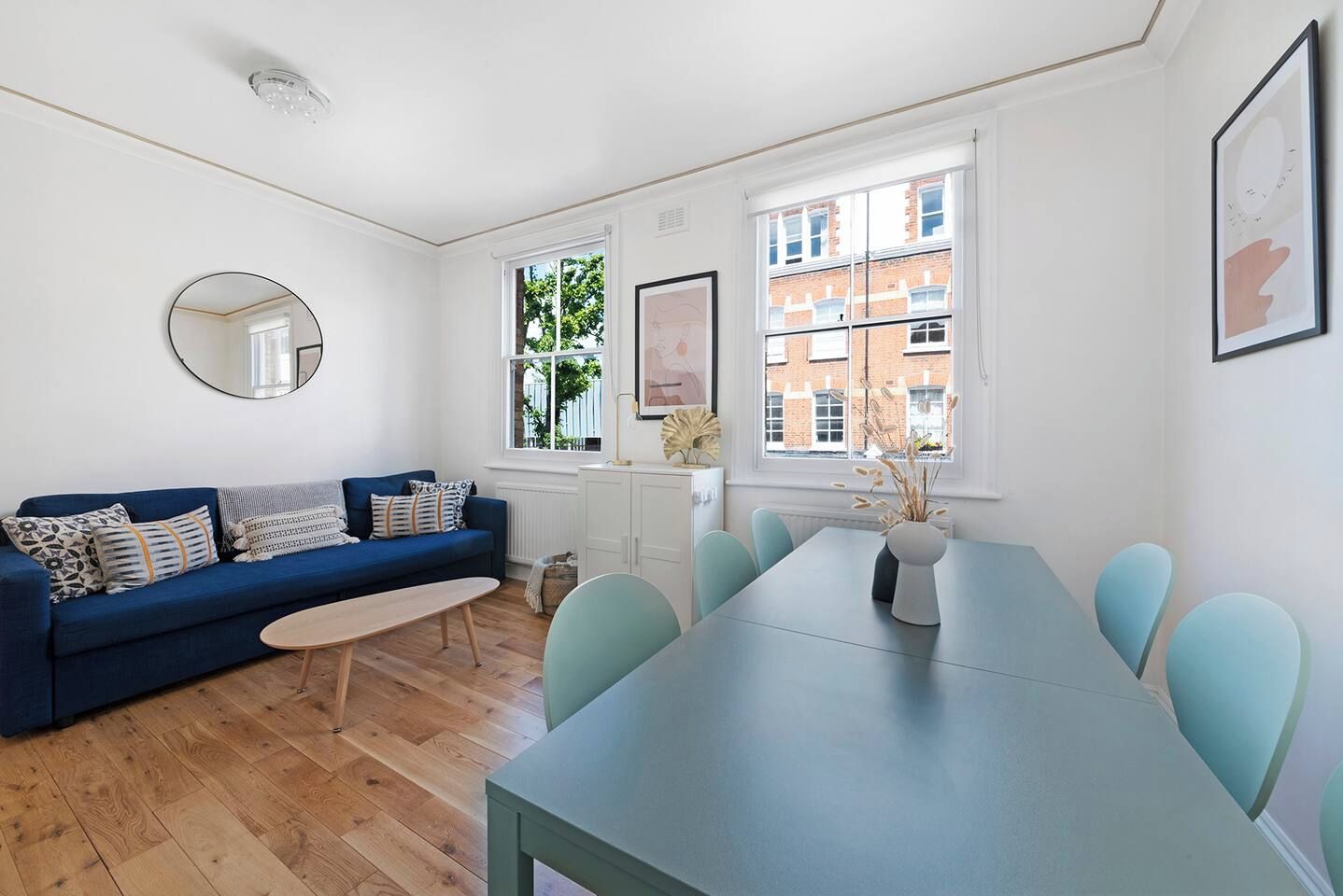 En moderne stue med blå sofa, rundt spejl, spisebord med stole og udsigt til en murstensbygning gennem store vinduer.