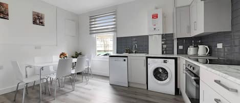 A modern kitchen with white cabinetry, a built-in washing machine, and a dining area with white chairs.