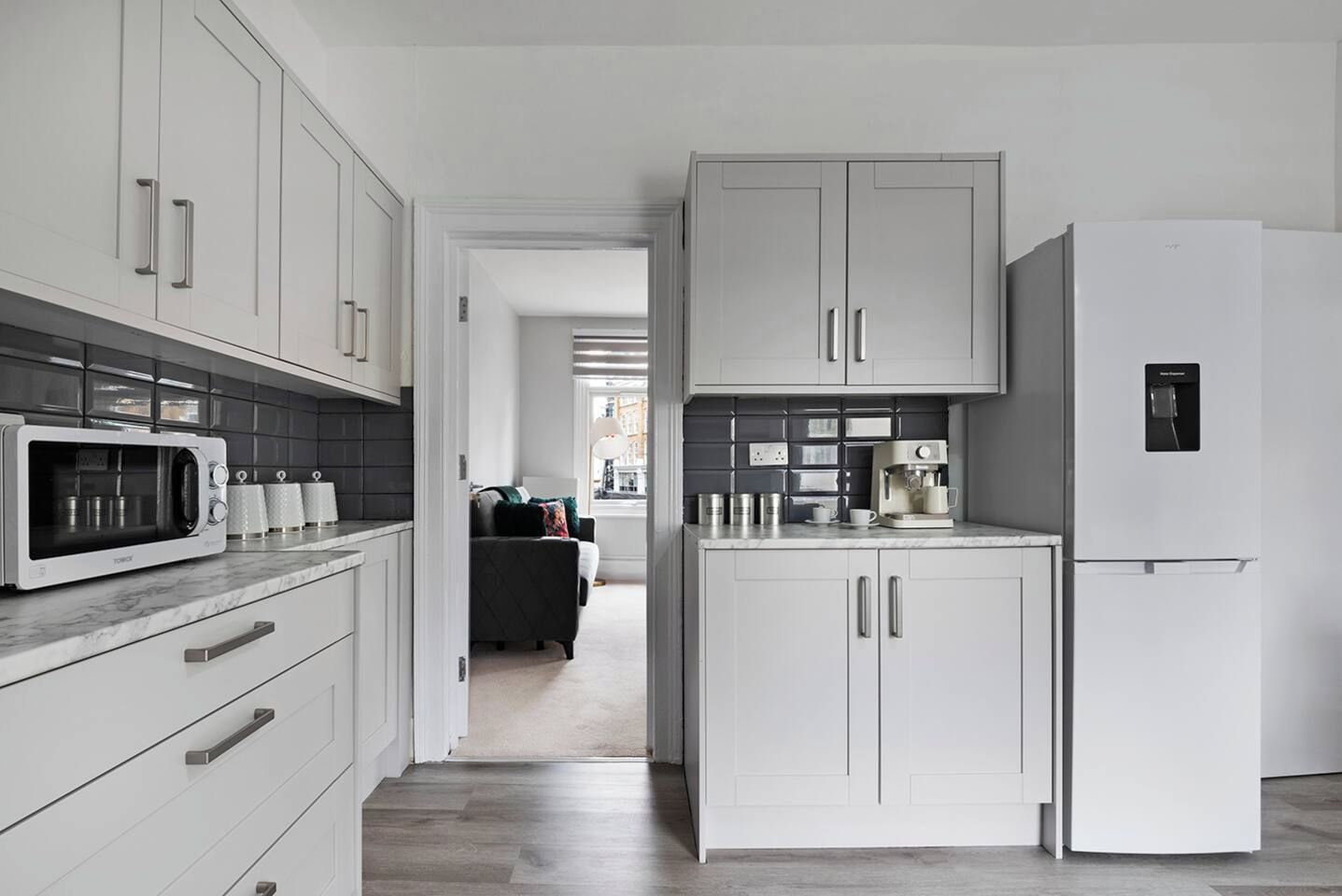 Una cocina moderna con armarios blancos, microondas, cafetera y refrigerador.