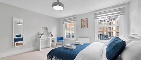 A bedroom with a bed, a desk, a chair, a mirror, and windows with a city view.