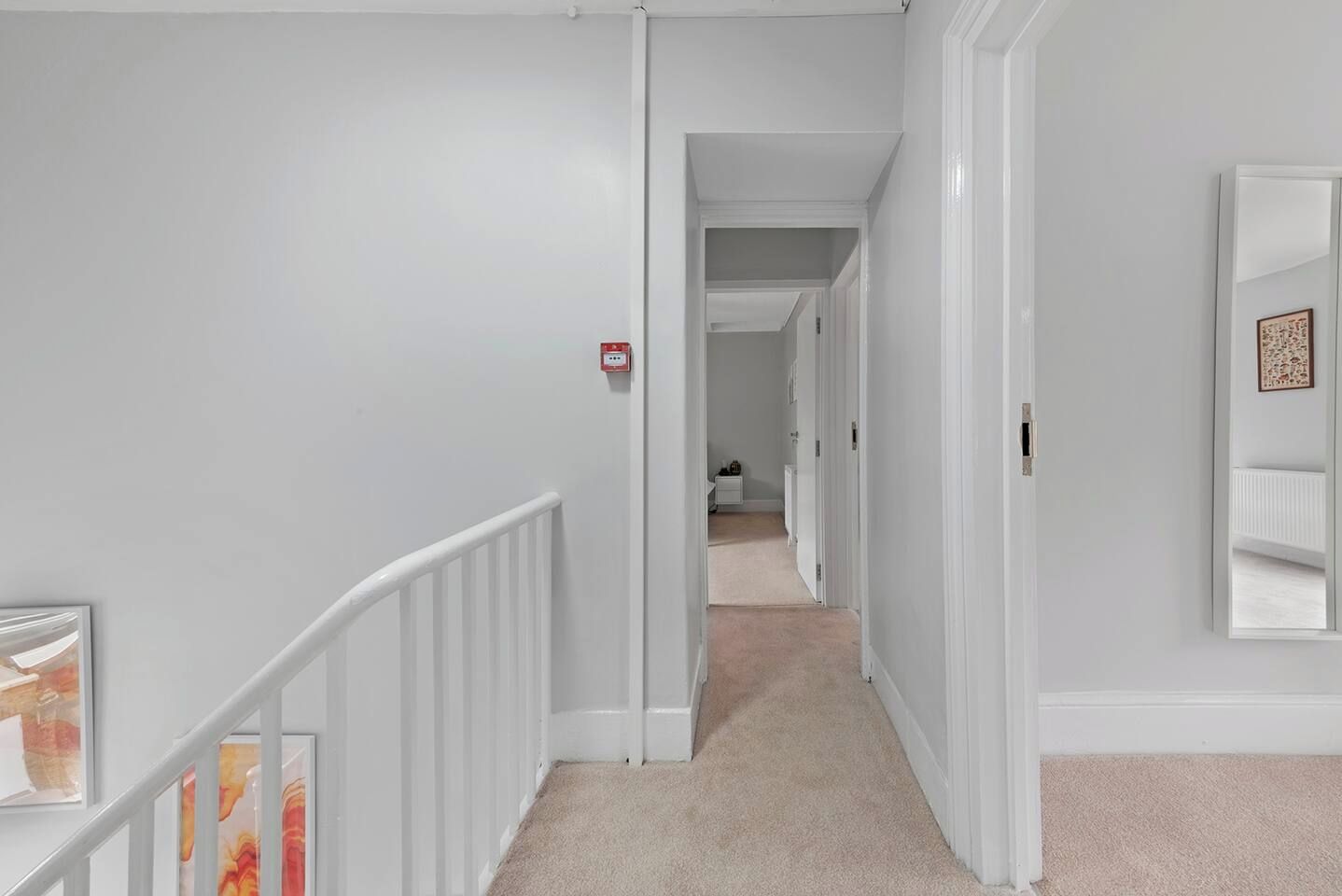 A hallway with a white railing, a fire alarm, and a carpeted floor leading to another room.