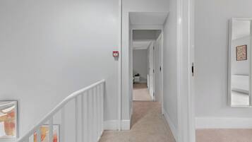 A hallway with a white railing, a fire alarm, and a carpeted floor leading to another room.