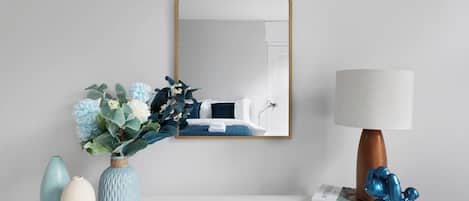 Une table de chevet blanche, ornée d’un miroir, d’une lampe et d’objets décoratifs, notamment un vase avec des fleurs et un livre.