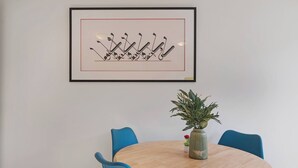 Ein Essbereich mit einem runden Tisch, blauen Stühlen, einem gerahmten Kunstwerk darüber und einer Topfpflanze auf dem Tisch.