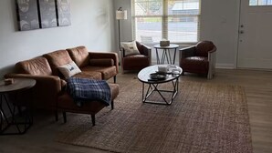 Ein Wohnzimmer mit braunem Ledersofa, einem Couchtisch und einem Teppich. Im Hintergrund befindet sich ein Fenster mit Jalousien und eine Tür.