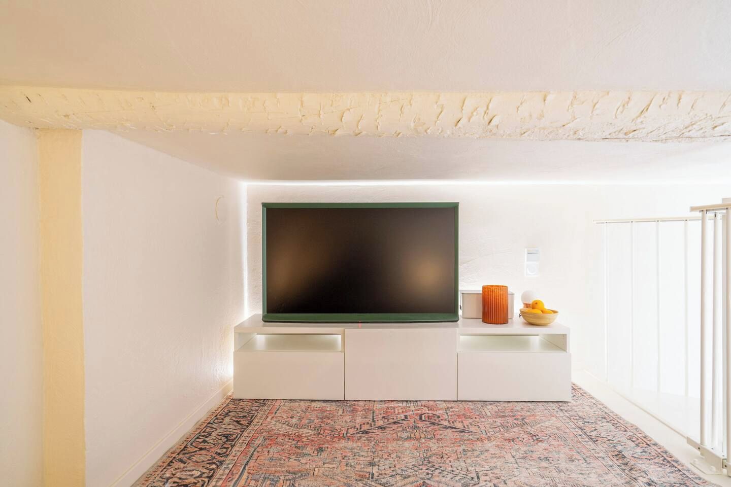 Una sala de estar moderna con un televisor de pantalla plana montado en un mueble blanco, un tapete estampado y una mesita con objetos decorativos.