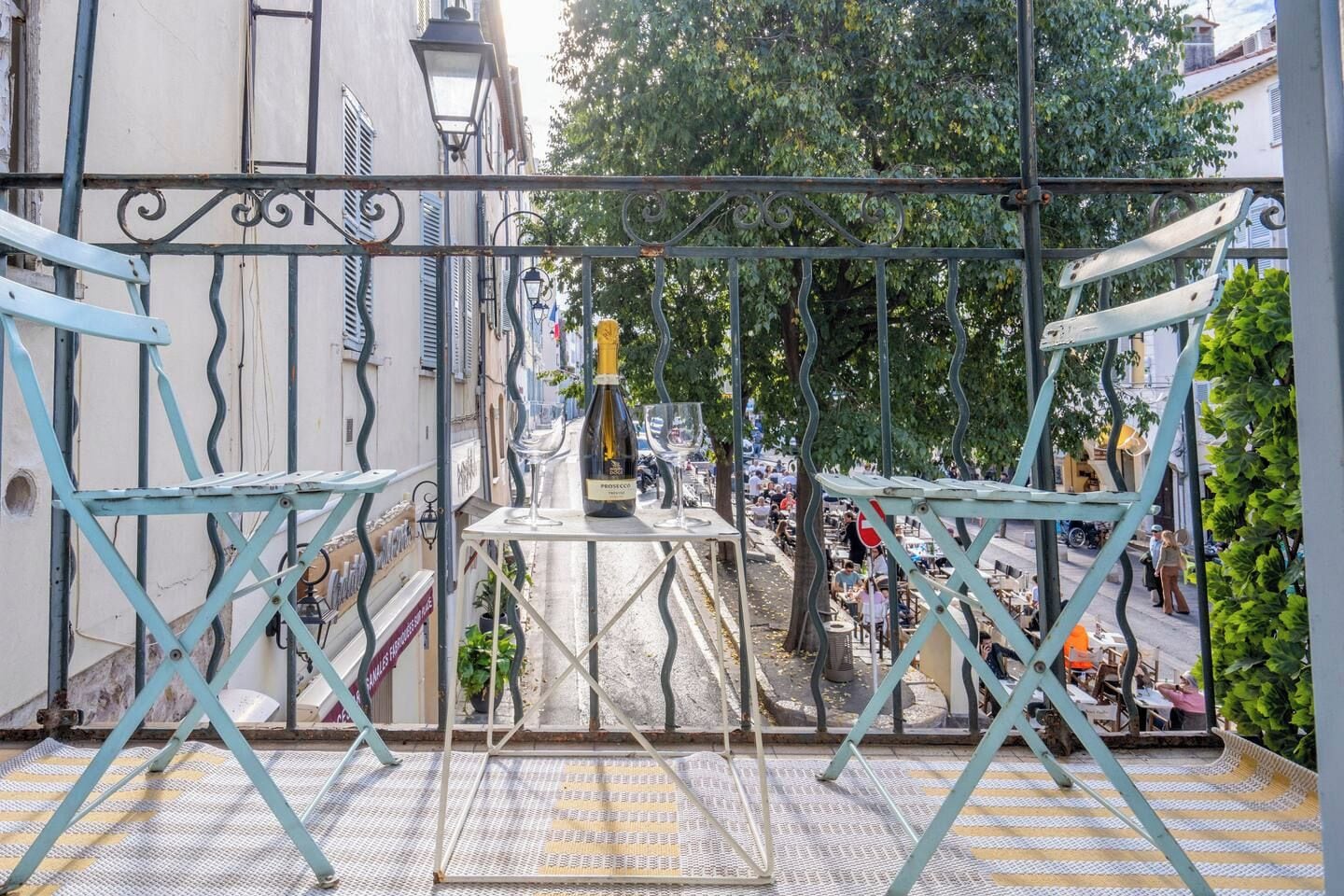 A balcony with a metal railing, a table with a bottle and glasses, and a view of a street with people and buildings.