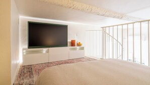 Ein Schlafzimmer mit einem Bett, einem an der Wand befestigten Fernseher, einem kleinen Tisch mit Schüssel und Kerze sowie einer Treppe, die zu einer oberen Etage führt.