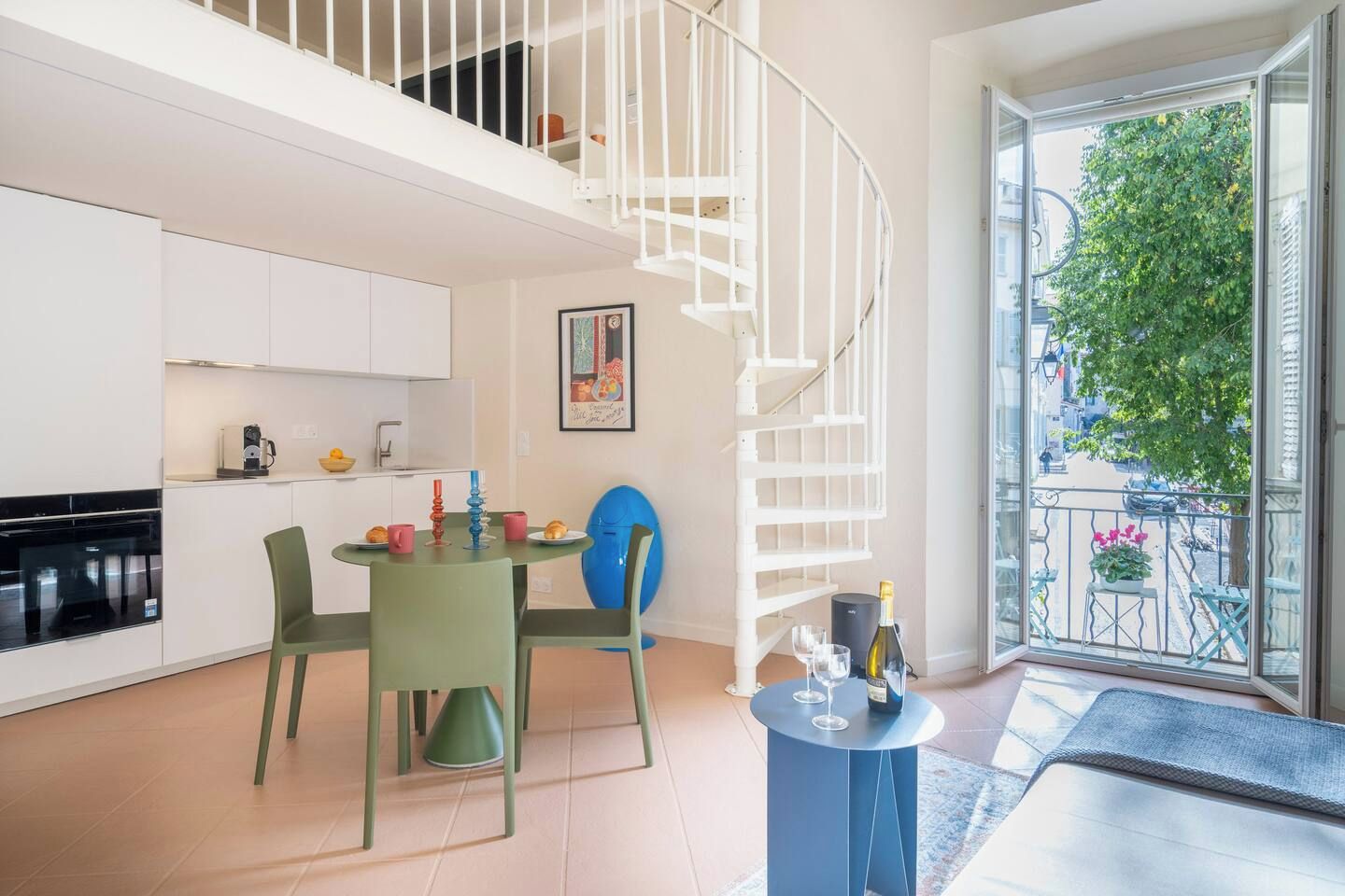 A modern kitchen with white cabinets, a dining area with green chairs, a blue table, and a spiral staircase.