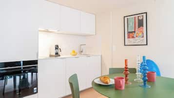 Una cocina compacta con armarios blancos, un horno negro, una cafetera y un lavamanos pequeño. Hay una mesa de comedor con un plato de comida y una taza rosa.