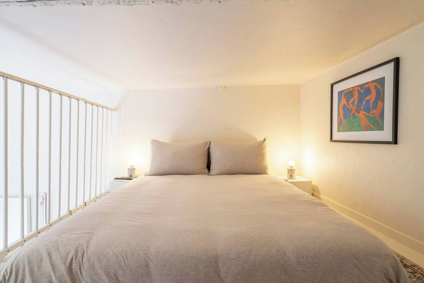 Un dormitorio con una cama grande, dos mesitas de noche, una mesilla auxiliar, una obra de arte enmarcada en la pared y una ventana con persianas.
