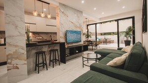 Ein modernes Wohnzimmer mit einem grünen Sofa, einem Couchtisch und einem an der Wand montierten Fernseher. Eine Küchenzeile mit Marmorarbeitsplatte und Pendelleuchten ist ebenfalls vorhanden.