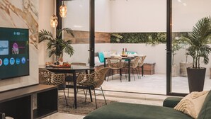 Ein modernes Wohnzimmer mit Flachbildfernseher, Korbmöbeln, einem Essbereich mit Tisch für zwei Personen und einem großen Fenster mit Blick ins Freie.