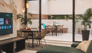 Una sala de estar moderna con una televisión de pantalla plana, mobiliario de mimbre, una zona de comedor con una mesa preparada para dos personas y un amplio ventanal con vistas al exterior.