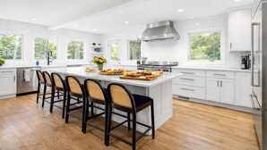 A modern kitchen with white cabinetry, a central island with bar stools, a stainless steel range hood, and a view of greenery outside.