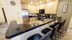 A modern kitchen with a granite countertop, stainless steel appliances, wooden cabinets, and a bar area with black barstools. - Remodeled Condo w/ FREE WiFi, Parking, Heated Pool, Hot Tubs, Skier Shuttles (Avon)