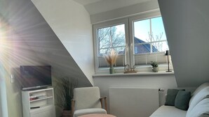 Un soggiorno moderno con un divano, un tavolo rotondo in legno, una TV su un mobile bianco e una finestra con vista su alberi e una casa.