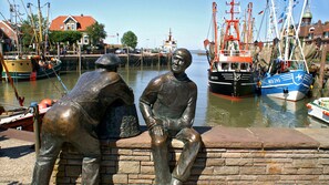 Eine Statue von zwei Personen, die auf einer Steinsitzbank an einem Yachthafen sitzen. Im Hintergrund sind Boote und Gebäude zu sehen.