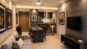 Una sala moderna con un sofá, un comedor con sillas, una cocina al fondo y un televisor de pantalla plana montado en la pared.