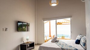 Ein Schlafzimmer mit Bett, einem an der Wand montierten Fernseher, einem Nachttisch mit Lampe und Blick auf den Balkon.