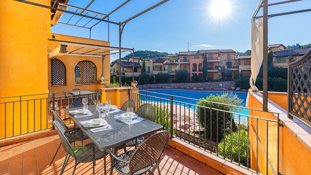 Un balcón con una mesa lista para comer, con vista a una piscina y una colina llena de edificios.