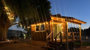 A wooden cabin with a deck, surrounded by trees and string lights. - Bob White Tiny Cabin (Quitaque)