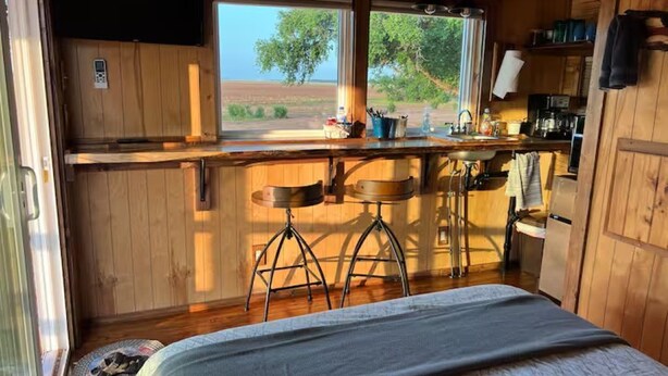 A compact kitchen with wooden cabinets, a bar stool, and a window overlooking a natural landscape. - Bob White Tiny Cabin (Quitaque)