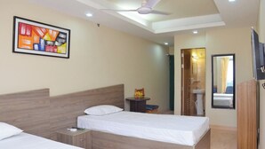 A hotel room with two beds, a ceiling fan, a framed abstract painting, a small table with fruit, a mirror, and a TV. - Hotel Titas View Brahmanbaria (Brahmanbaria Sadar)
