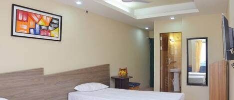 A hotel room with two beds, a ceiling fan, a framed abstract painting, a small table with fruit, a mirror, and a TV.