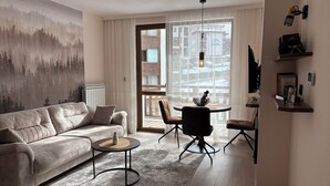 A modern living room with a beige sofa, a round coffee table, and a dining area with a small table and chairs. - ✨ Elegant Mountain Apartment with Fireplace, Balcony Views, SKI In&Out ⛷ (Bansko)