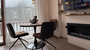 A modern living room with a fireplace, a dining area with two chairs, a TV mounted on the wall, and a view of the outdoors through a large window. - ✨ Elegant Mountain Apartment with Fireplace, Balcony Views, SKI In&Out ⛷ (Bansko)