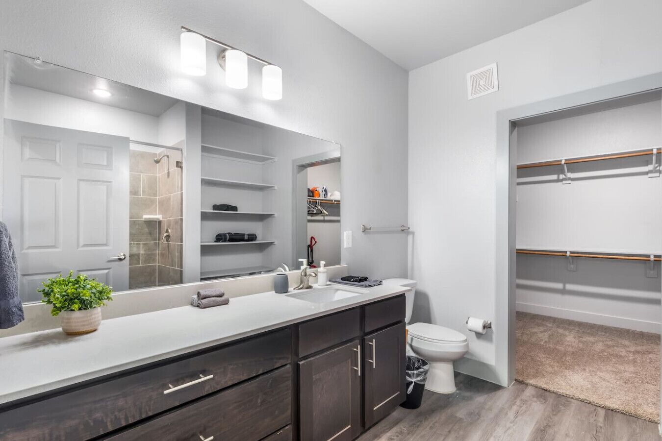 Une salle de bain moderne dotée d’un grand miroir, d’un meuble-lavabo avec des placards de couleur sombre, de toilettes et d’un dressing avec espace pour suspendre les vêtements.