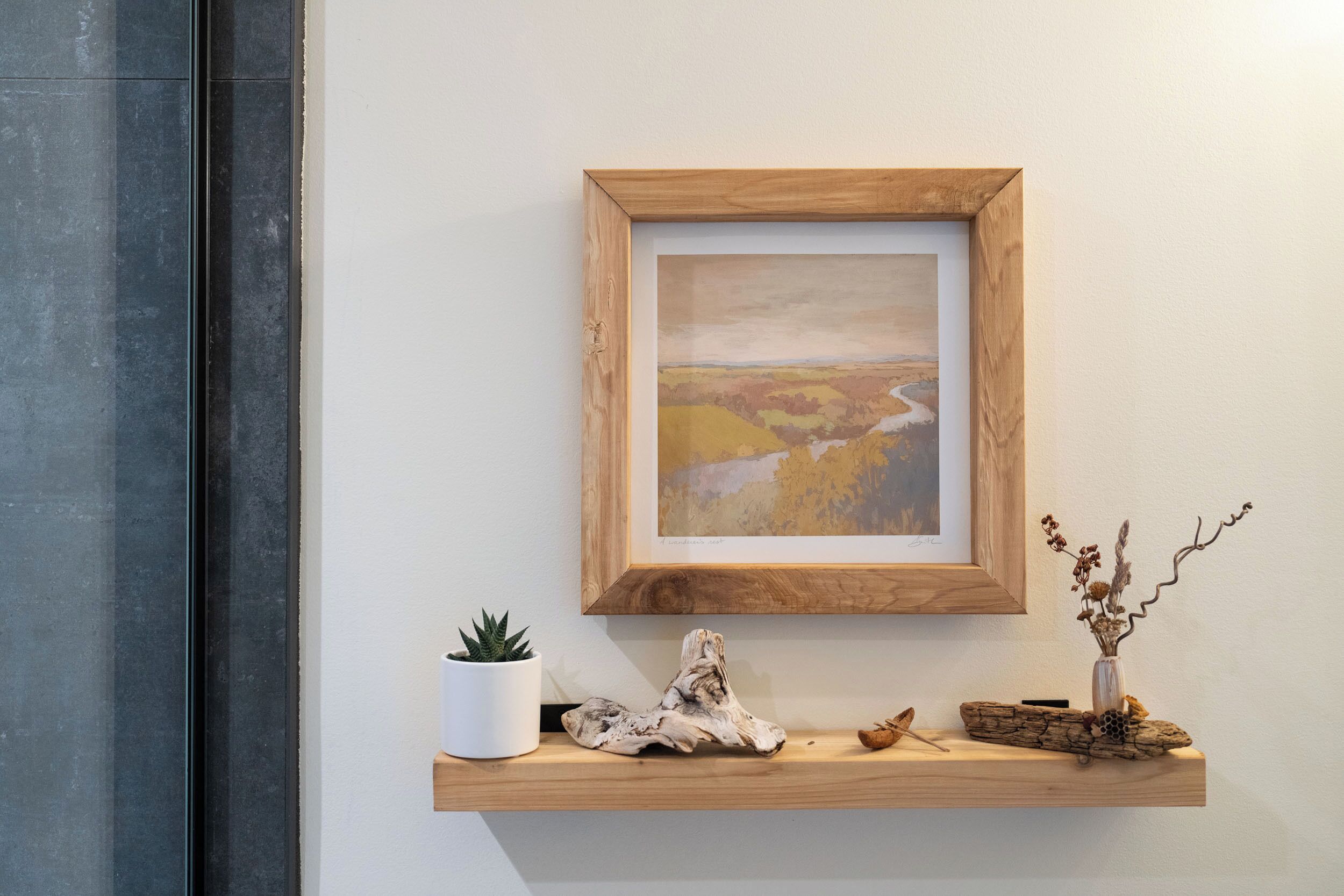 Un tableau représentant un paysage, encadré et accroché au mur, une petite plante en pot et un objet décoratif en bois posé sur une étagère.