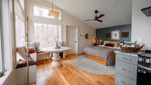 A spacious bedroom with a large bed, a seating area with a round table, and a view of trees through the windows. - the Birdhouse of Boone, Luxe + Long Views + Charming Tiny Home (Boone)