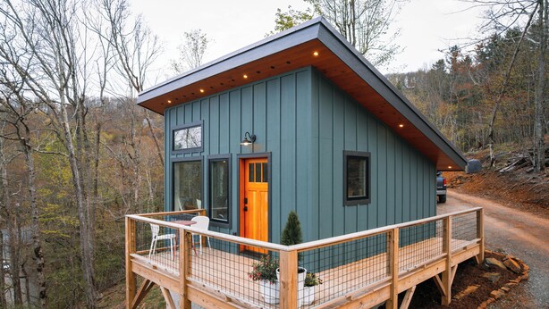 A wooden deck with a railing, a small potted plant, and a door with a light fixture. - the Birdhouse of Boone, Luxe + Long Views + Charming Tiny Home (Boone)