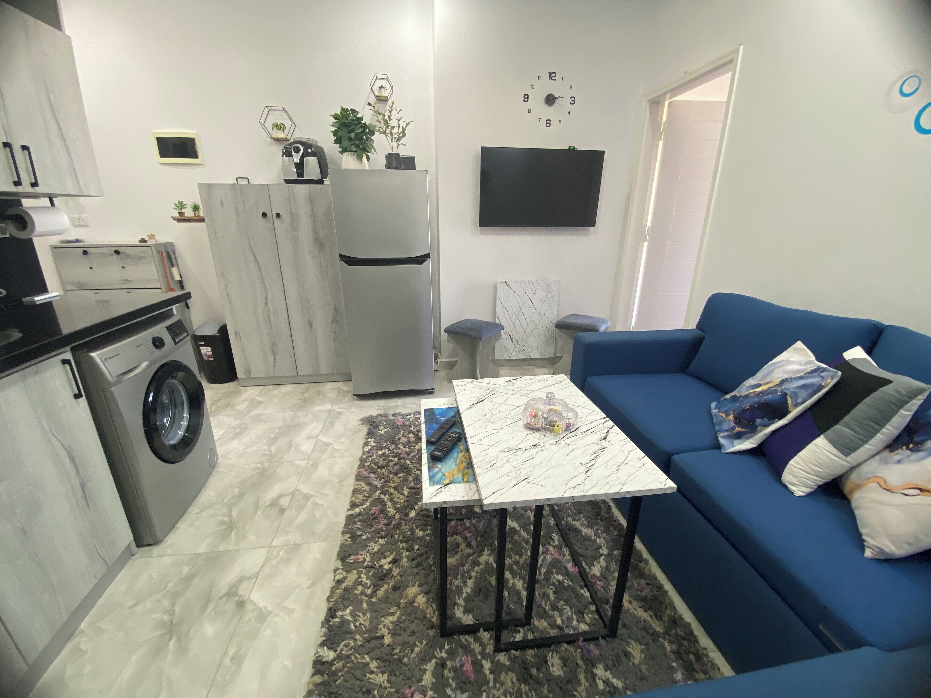 A compact living space with a washing machine, a refrigerator, a sofa, a coffee table, and a TV mounted on the wall.