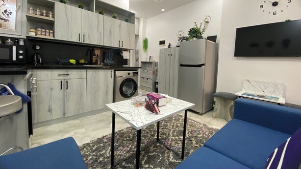 Een moderne keuken met grijze kastjes, een wasmachine en een koelkast. Er staat een eettafel met een vaas en een schilderij hangt aan de muur.