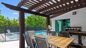 Een terras buiten met een zwembad, een houten tafel, stoelen en een barbecue.