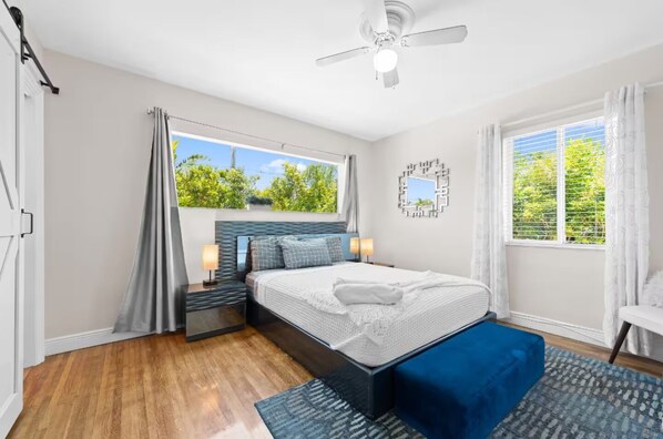 A modern bedroom with a large bed, a ceiling fan, a window with curtains, and a view of greenery outside.