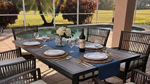 Un espacio de comedor al aire libre con una mesa dispuesta para seis personas, rodeado de sillas de mimbre, con vistas a una piscina y palmeras.