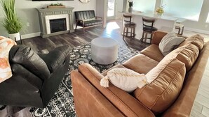 A modern living room with a brown sofa, a black leather chair, a patterned rug, a fireplace, a flat-screen TV, and a dining area with a table and chairs. - LAKE THERAPY- Cozy Lakeside with Beautiful Sunsets! (Temple)