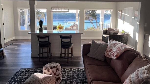 A modern living room with a brown sofa, a patterned rug, and a view of a lake through large windows. - LAKE THERAPY- Cozy Lakeside with Beautiful Sunsets! (Temple)
