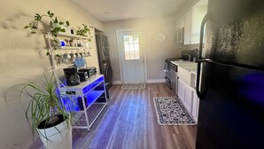 Eine kompakte Küche mit Kühlschrank, Backofen und Mikrowelle. Es gibt ein kleines Spülbecken und eine Arbeitsplatte mit Schränken. Eine Topfpflanze befindet sich links, und eine blaue Regalwand ist an der Wand angebracht.
