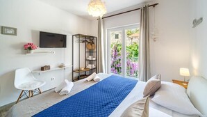 Ein Hotelzimmer mit Bett, Fernseher, Sessel, Ablagefläche mit Handtüchern und Balkon mit Blumen.