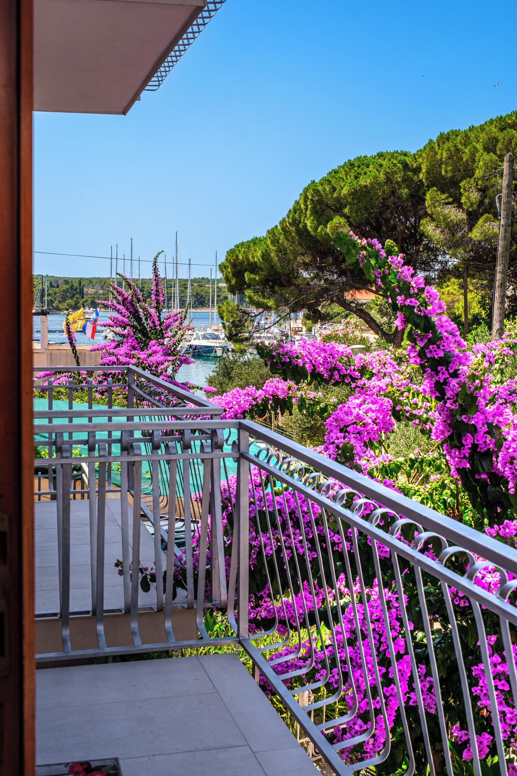 Un balcon doté d’une balustrade en métal, donnant sur une marina où sont amarrés des bateaux, avec un ciel bleu dégagé.