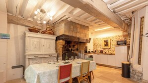 A dining area with a table set for four, a stone fireplace, and a kitchen in the background. - Maison 1505 | Authentic 16th-century shipowner’s house in the city center (Honfleur)