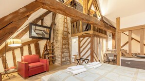 A room with exposed wooden beams, a red sofa, a bed with a patterned bedspread, a ladder, and a small table with a lamp. - Maison 1505 | Authentic 16th-century shipowner’s house in the city center (Honfleur)