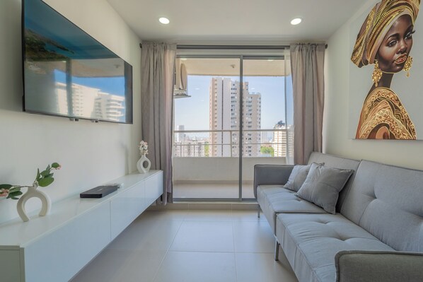 A modern living room with a grey sofa, a flat-screen TV mounted on the wall, a balcony with a view of high-rise buildings, and a decorative painting on the wall. - Very close to the beach and the historic center (Cartagena de Indias)